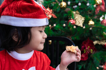 A cute Asian little girl in a red Santa Claus dress  eating bread ​on Christmas eve. Christmas in COVID 19 Concept