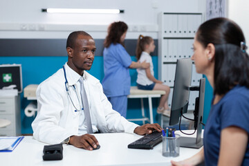 Fototapeta premium African american pediatrician doctor explaining disease symptoms to patient mother typing medical treatment on computer keyboard. Specialist man working in hospital office during clinical consultation