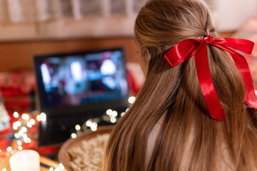 behind female head with long blondie hair and red bow rear view watching the christmas movie on laptop at camping home in cozy bed with glowing lights garland at night on christmas or new year eve