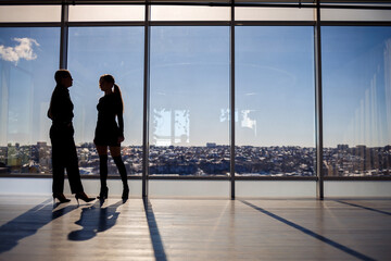 Two business women enjoying the city view and talking while standing by the large window in the office