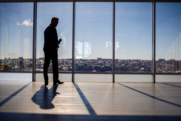 Fototapeta na wymiar Rear view of a businessman looking out of a large window overlooking the city. He has a phone in his hands. Horizontal view. Selective focus