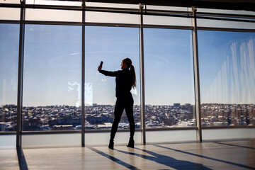 Fototapeta na wymiar A beautiful girl takes a selfie on her phone against the background of panoramic windows in a skyscraper. Selective focus