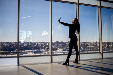 Fototapeta na wymiar A beautiful girl takes a selfie on her phone against the background of panoramic windows in a skyscraper