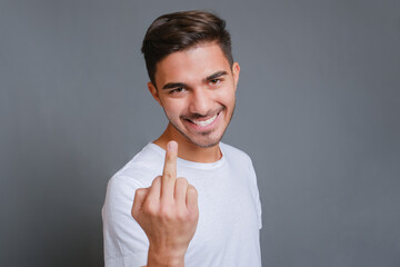 Close-up young smiling caucasian man wearing white t-shirt standing over isolated gray background and showing middle finger