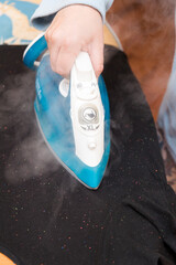 Vertical photo of a woman's hand ironing a black T-shirt on an ironing board, steam coming out of the iron.