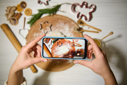 Top view of hands holding mobile phone and making photo of a Gingerbread dough on a round wooden board next to a rolling pin and kitchen utensils for making Christmas cookies. Phone in live view mode