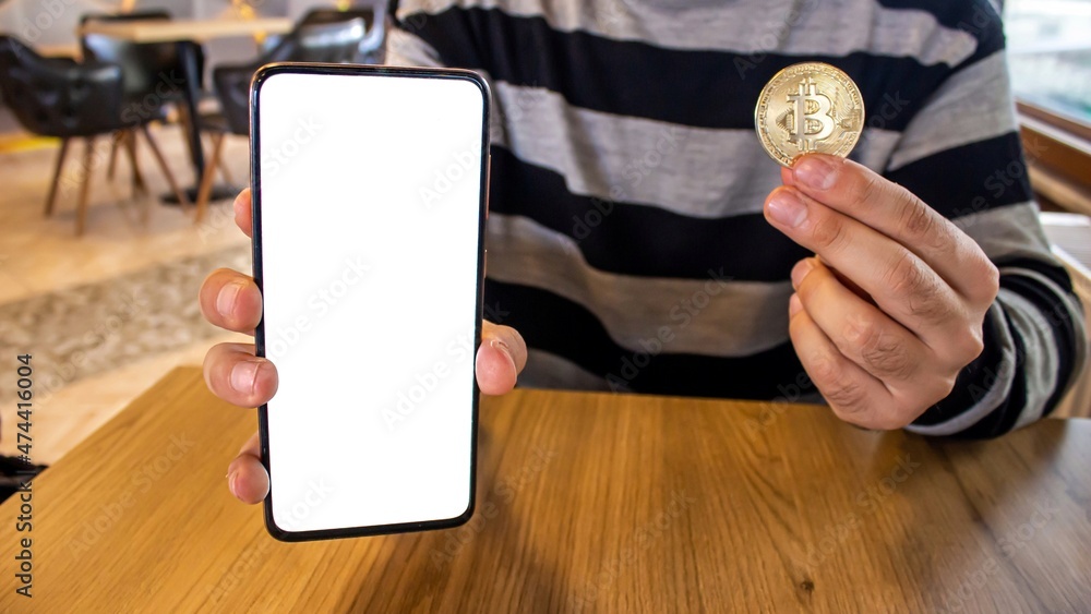 Poster man holding bitcoin coin and showing mobile phone with blank white screen while sitting in a cafe.