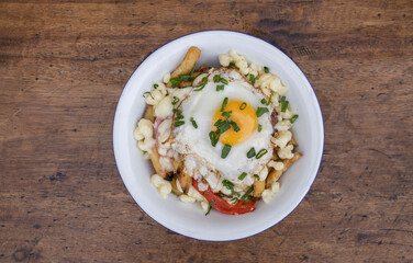 Top view of fried potatoes with melted cheese, chorizo, bell pepper, onion and a grilled egg in a white bowl on the wooden table.  