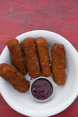 Finger food. Top view of fried breaded mozzarella sticks with dipping barbecue sauce in a white bowl on the red wooden table. 
