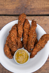 Fried gourmet food. Overhead view of deep fried chicken sticks breaded with seeds and a dip made of mustard and honey, in a white bowl on the table.