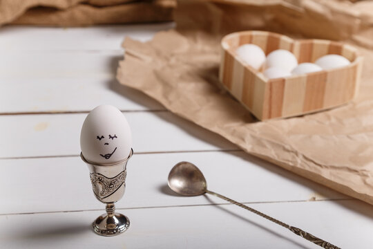 Easter egg with painted face in silver egg cup with spoon on white wooden table. Unfocused heart shaped box with eggs on background. Easter concept.