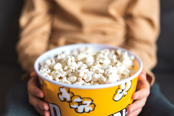 From above of crop unrecognizable person sitting on sofa with bucket full of yummy popcorn