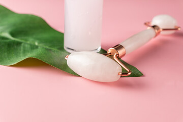 Face roller and serum dropper bottle on pink background with green leaf. Home spa procedures.