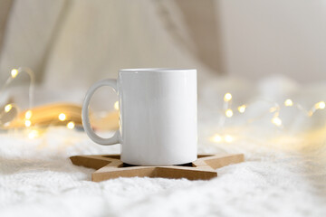 Obraz na płótnie Canvas White mug mockup with blank copy space. Coffee cup on a beige knited background with opened book and christmas garland lights, cozy winter day at hugge home