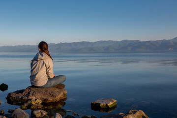 Fototapeta premium Quiet sunset over the river. The girl is sitting on a big stone, summer calm evening.