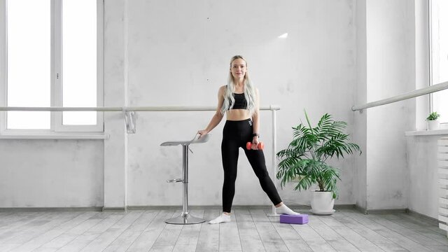 Woman doing exercise for legs and arms. Sporty blonde training at home using dumbbell. Healthy lifestyle. Young woman doing squats in black leggings indoors. 4K, UHD