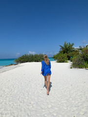 A back view of a tanned long-haired girl in a short blue dress running along the shores of the Indian Ocean with white sand and azure water in the Maldives.