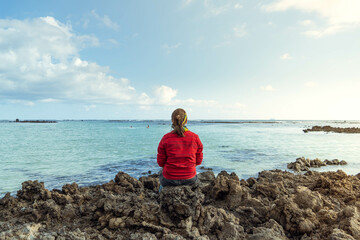 Mujer joven de espaldas con chaqueta roja sedada sobre unas rocas volc&aacute;nicas admirando el paisaje mar&iacute;timo.