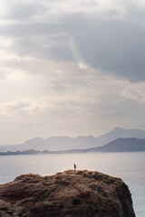 Fotografía vertical de un hombre joven en una roca con el mar y una isla montañosa detrás. - obrazy, fototapety, plakaty
