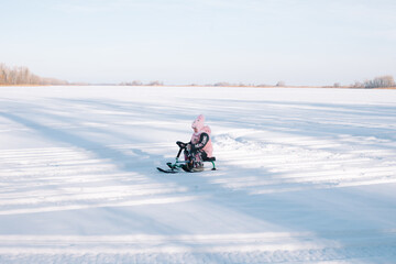 Child rides snowcat on snowy road. Little girl in pink warm jacket enjoys walk in nature and sledding on frozen river on sunny winter day, side view. 