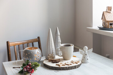 A cup of coffee on a wooden stand, ceramic Christmas trees in pastel colors, gingerbread cookies, gray candlestick. Artificial spruce branch with red berries. Scandinavian style. Copy space. - obrazy, fototapety, plakaty