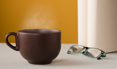 Brown cup of coffee or tea with hot steam, on the table, with books, glasses and brown background.
