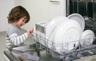 Child helping to unload dishwasher with white dishes. Girl helping in the kitchen. Child making...