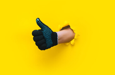 A man's hand in a black fabric work glove shows a thumb up (like). Torn hole in yellow paper. Good...