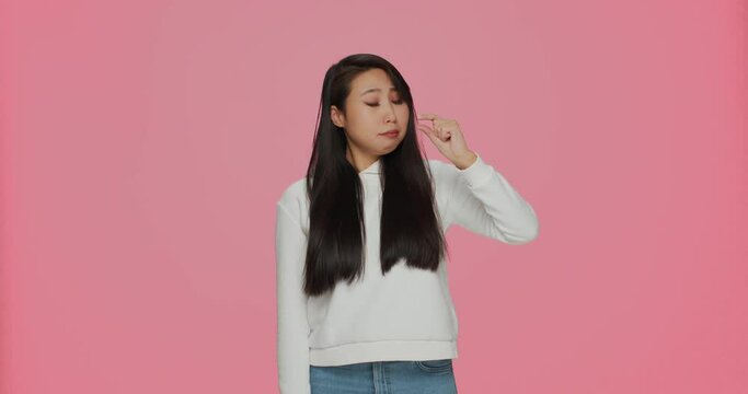 Upset young asian girl showing with fingers small size gesture, little measurement on pink studio background