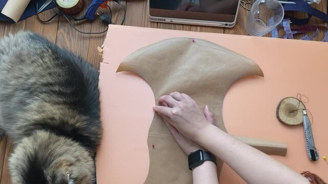 Women's hands attach sheet of paper in form of viking axe to piece of styrofoam with pins. Fluffy cat lies on wooden table in cosplayer's workshop.