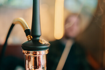 Hookah on table in lounge bar. Smoking person in blurry background. Close-up of black pipe and golden shisha flask. Leisure in oriental cafe.