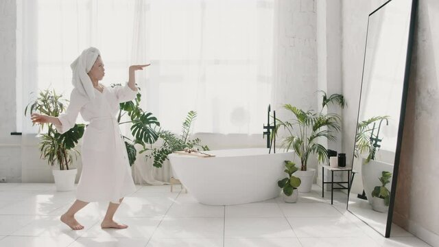 Young caucasian woman in white bathrobe and towel on her head dancing with glass of champagne after morning shower at home, slow motion. People, joy and music concept. Woman alone happiness concept