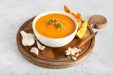 Wooden board with bowl of tasty pumpkin cream soup, seeds and garlic on light background