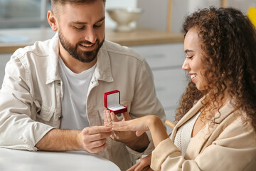 Young man putting engagement ring on his girlfriend's finger at home