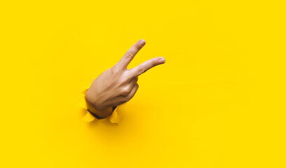 Victory sign two-finger hand gesture on yellow background with torn paper hole and copy space. It...