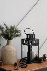 Black Christmas lantern with burning candle, vase, fir branches and cones on table near light wall