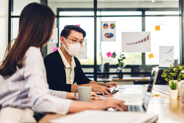 Asian business man and woman using laptop computer working and planning meeting in quarantine for coronavirus wearing protective mask with social distancing while sitting on office desk