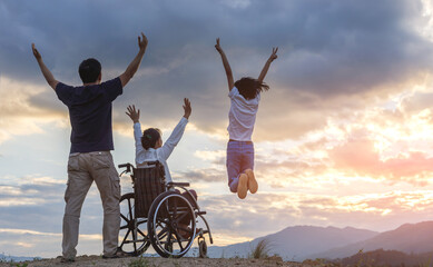 Group of Happy Family with Woman Sitting in wheelchair raised hands on mountain at sunset background.