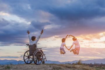 Woman Sitting in wheelchair raised hands and children jumping on mountain at sunset background....