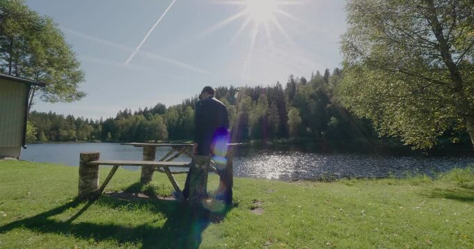 Young Man With Cap and Bomber Jacket Sits at Bench at Kypesj&ouml;n Lake in Bor&aring;s Sweden - Wide Shot Static