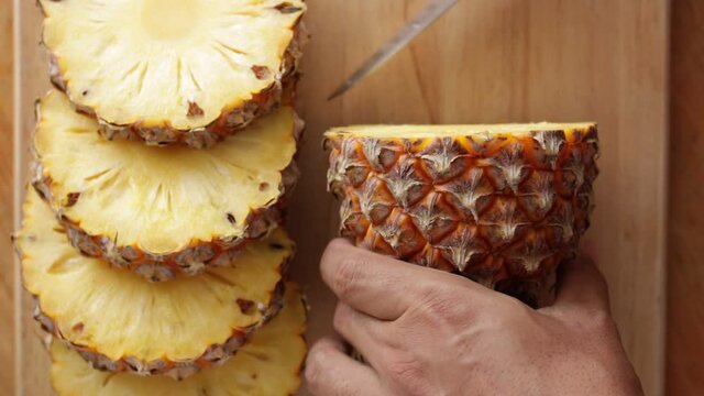 CU Close-up of Man's hand slicing pineapple with a knife on a wooden cutting board. 