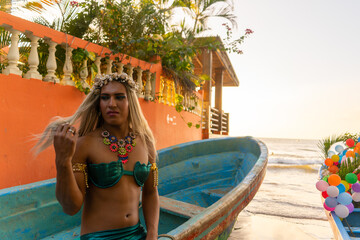 Diverse Latino gay man dressed as a mermaid with long blonde hair and ornaments on his hair and neck,fixing hair on the beach at sunset taking pride in being a part of the lgbtq community