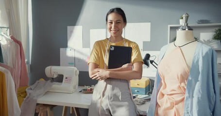 Cheerful professional Asia lady fashion designer with casual hold digital tablet smile look at camera with confidence in her shop. Lady tailor student proud of her new collection in workshop studio.