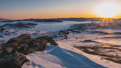 Winter dawn in Siberia. The sky is highlighted in orange. Glare from the sun's rays on the ice of a frozen lake. Campsite on the shore. In the foreground is a rock covered with sparkling snow. Baikal