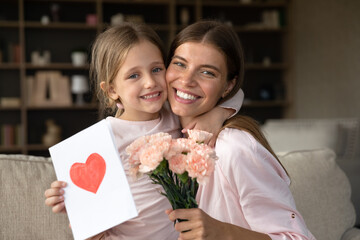 Grateful mom hugging daughter girl, holding flowers bouquet, receiving hand drawn greeting card with loving heart from girl, smiling at camera. Mothers day, 8 march, concept. Head shot portrait