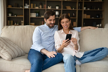 Happy millennial couple using online shopping app, internet service, making video phone call, sharing cellphone, watching media content together, talking, resting on sofa with digital device at home