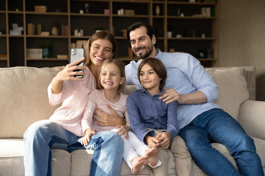 Happy excited parents, little son and daughter resting on couch, using digital gadget, taking selfie on mobile phone, recording video for blog, looking at smartphone screen, smiling, laughing