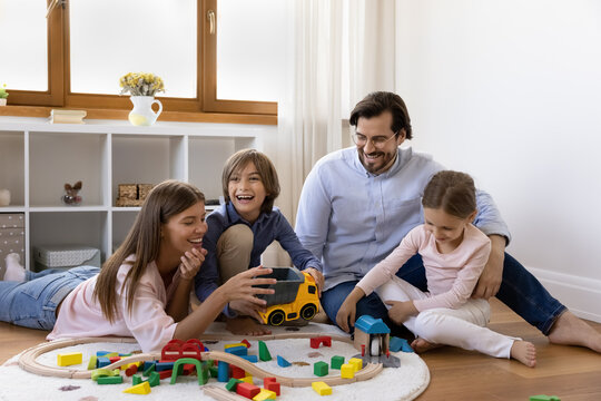 Happy caring parents and little children playing games on warm floor at home together, driving toy truck, train on railway, constructing town from colorful building blocks, having fun with family