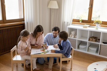 Mom and dad teaching little kids to draw in colored pencils, helping sibling children with school homework task, sitting at small wooden desk together. Family home activities, craft hobby concept