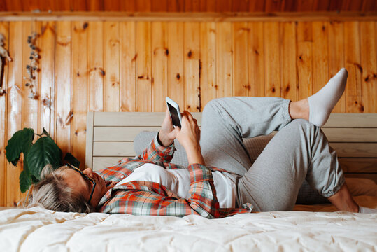 Adult woman chatting, browsing social media on mobile phone while relaxing, resting on bed in bedroom. Side view of lying caucasian woman in casual clothes using smartphone